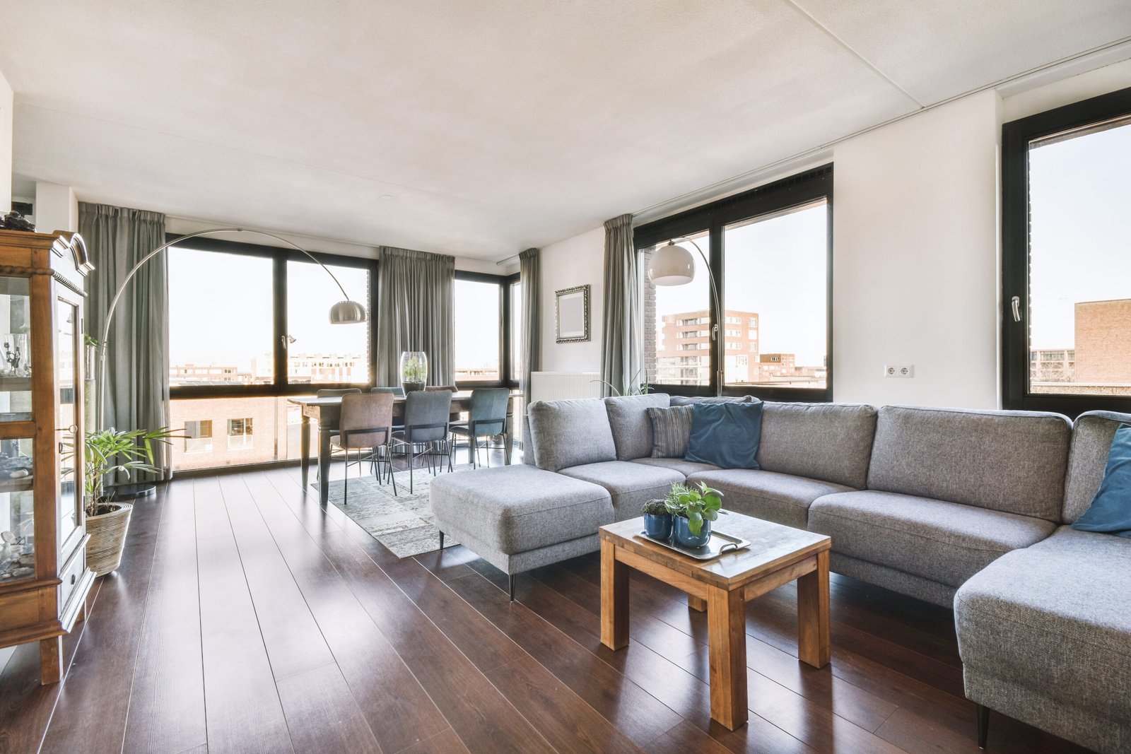 Wooden table with chairs and vase located near comfortable couch against light walls large windows in modern apartment