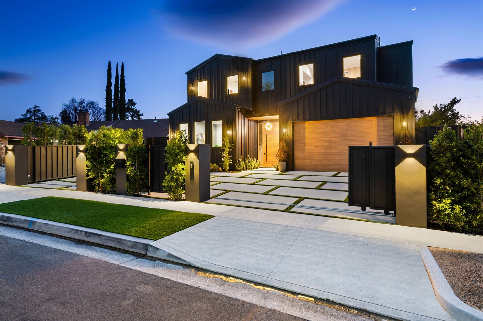 Modern luxury house exterior with well-lit driveway and lush landscaping during twilight.